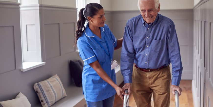 Senior Man At Home Using Walking Frame Being Helped By Female Care Worker In Uniform