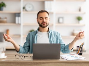 Workplace stress management. Calm Caucasian man meditating with closed eyes in front of laptop pc at home office